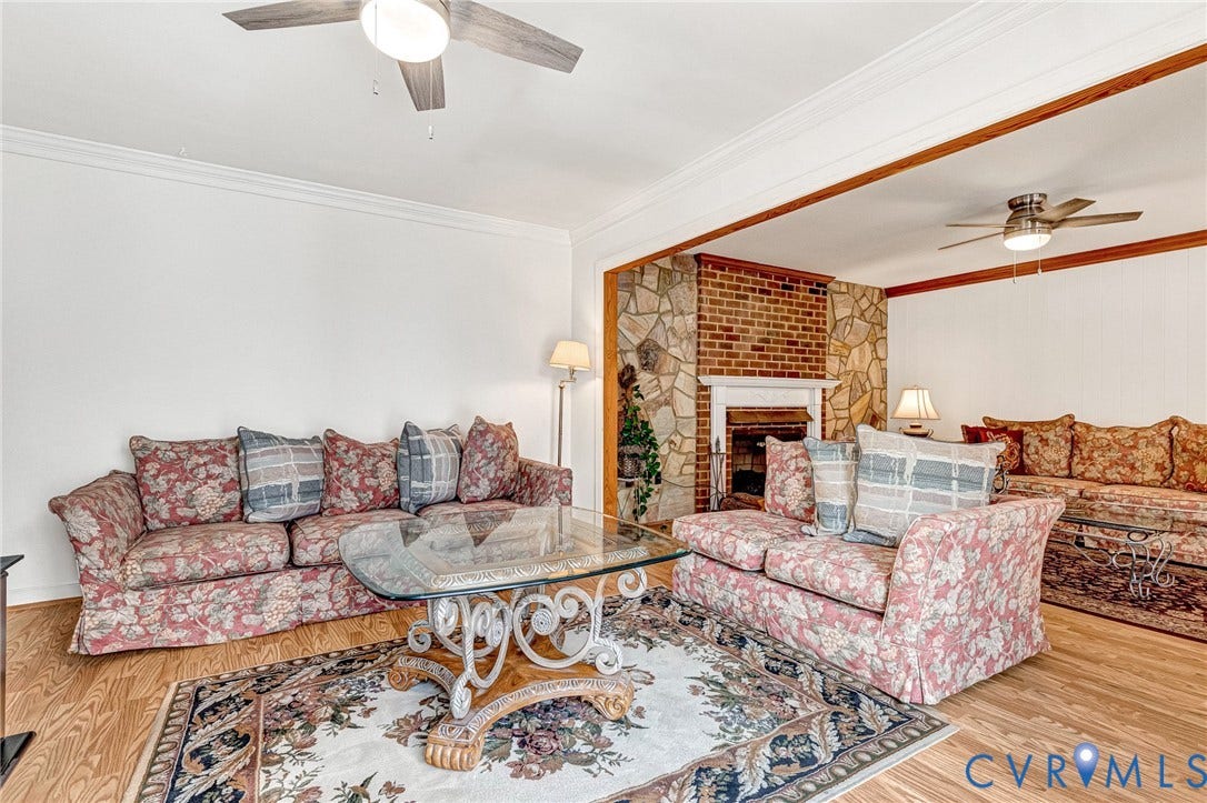 Living area featuring wood-finish flooring, a stone and brick fireplace, crown molding, and two ceiling fans