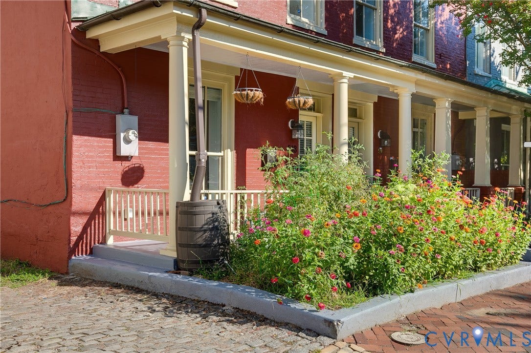 518 N 1st Street, Jackson Ward, Richmond, VA 23219 Red brick exterior with white trim, featuring a covered porch with white columns, a garden bed with flowering plants, a white porch railing, and a cobblestone-style pathway