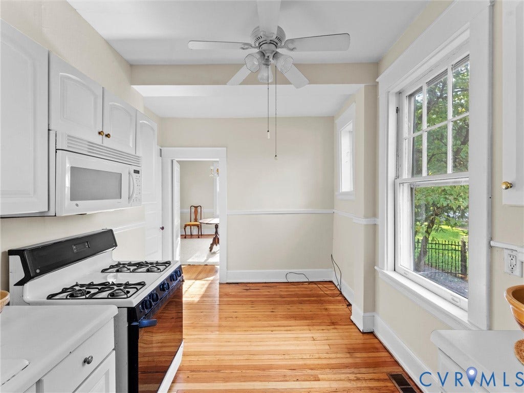 Galley kitchen showcasing white cabinetry with hardware accents, dual gas cooktops, stainless steel microwave, warm wood-finish flooring, and abundant natural light from multi-pane windows overlooking the grounds