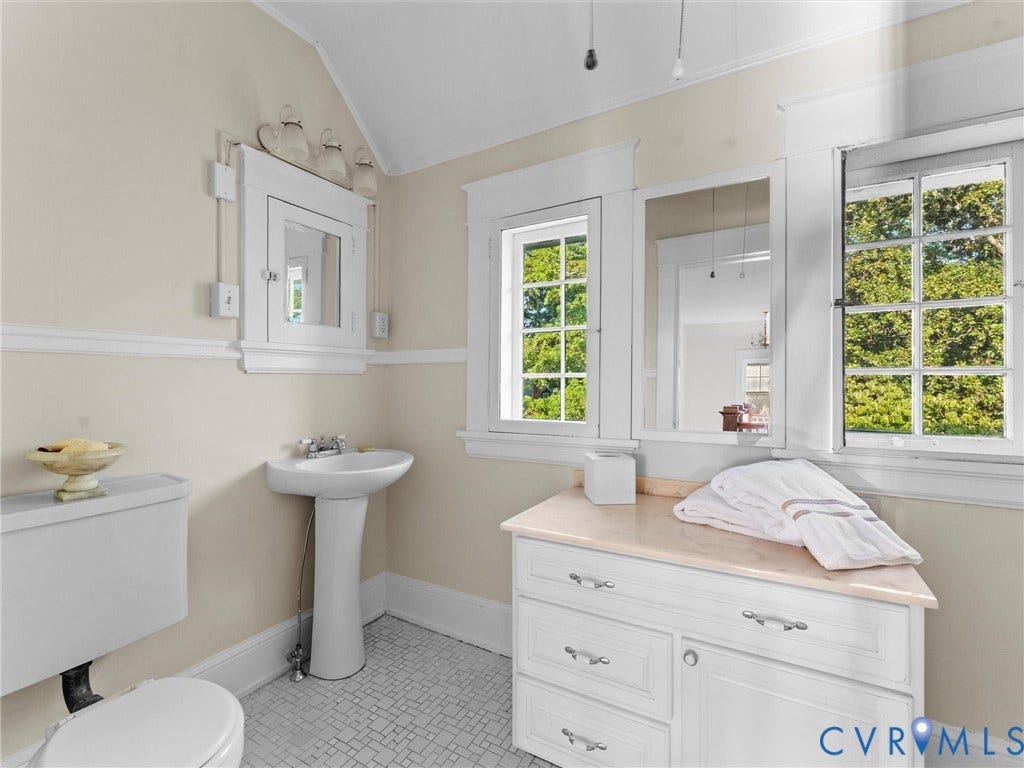 Bright, airy bathroom featuring a pedestal sink, white cabinetry with multiple drawers, hexagonal tile flooring, and abundant natural light from multi-pane windows