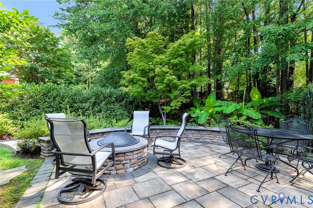 Paver patio featuring a stone-clad fire pit, low stone retaining wall, and lush perimeter landscaping