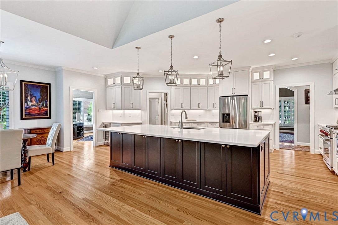 Spacious kitchen featuring hardwood flooring, a large island with a light-toned countertop, white cabinetry with under-cabinet lighting, stainless steel appliances, and decorative pendant lighting