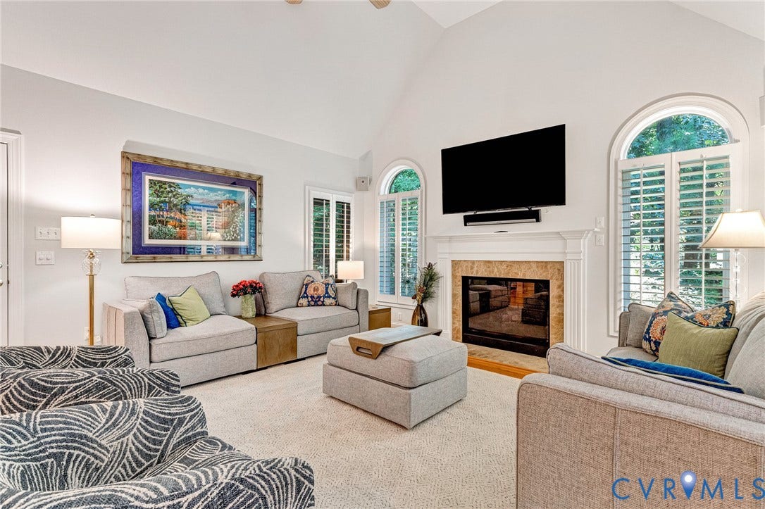 Vaulted ceiling, arched windows with plantation shutters, fireplace with a stone surround and mantle, and light-toned wall finishes