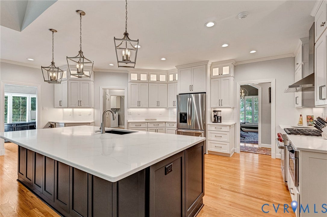 Spacious kitchen featuring a large central island with a white countertop, light wood-finish flooring, white cabinetry with upper glass-front elements, and stainless steel appliances