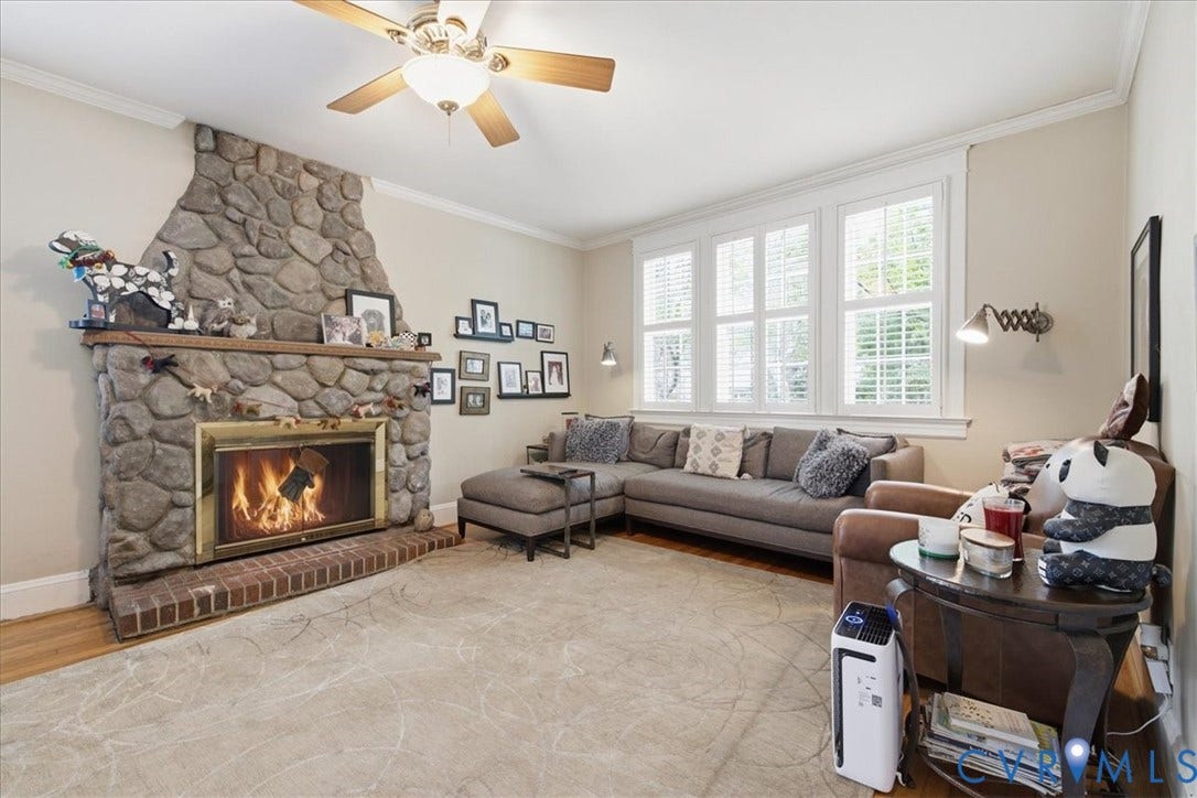 4702 New Kent Avenue, Westover Hills, Richmond, VA 23225 Stone look-clad fireplace with brick hearth, multiple transom windows with plantation shutters, crown molding, and a ceiling fan with integrated lighting