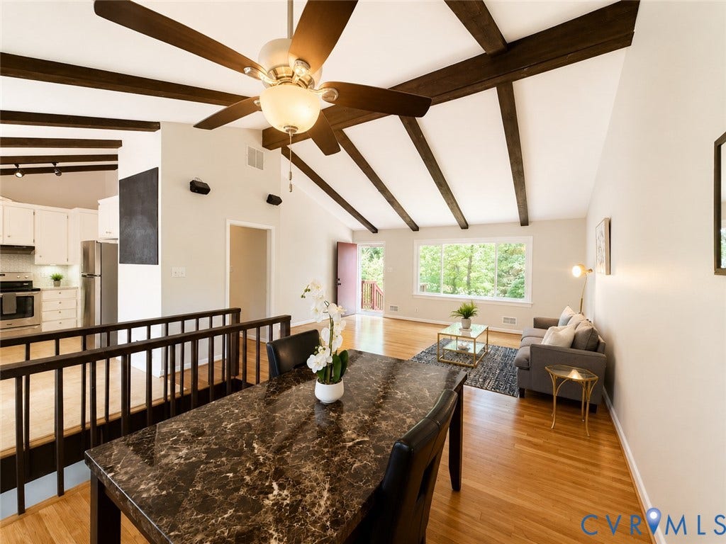 9018 Brucewood Drive, Midlothian, VA 23235 Open-concept living space featuring exposed wood beam ceiling with integrated ceiling fan, expansive windows with natural light, warm wood-finish flooring, dark stone countertop island, and black metal railing detail
