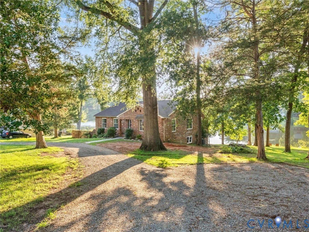 Brick exterior residence with a gravel driveway, surrounded by mature trees, and a body of water in the background