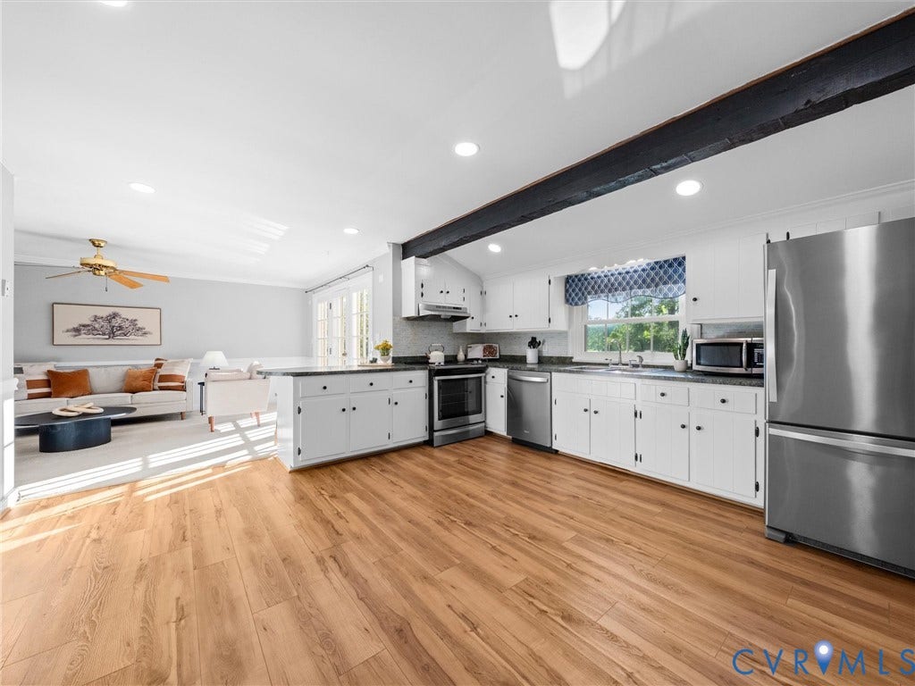 Wood-finish flooring throughout, white cabinetry with dark hardware, stainless steel appliances, dark countertops, and a wood beam accent