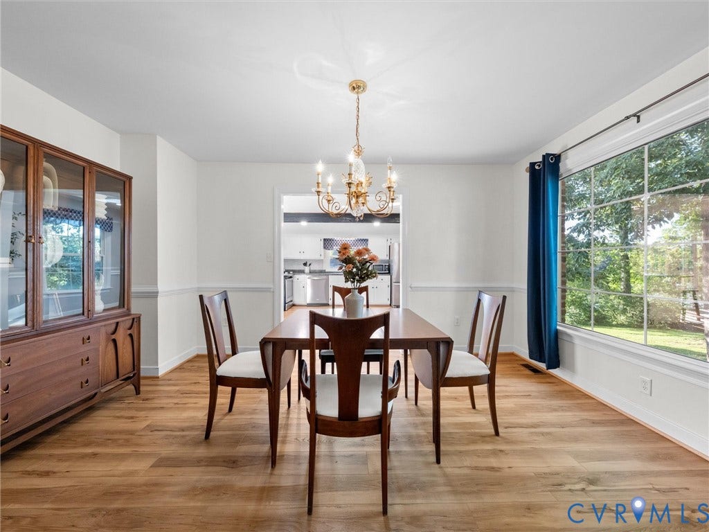 Wood-finish flooring, white walls with wainscoting, an ornate brass chandelier, and a large window with exterior scenery