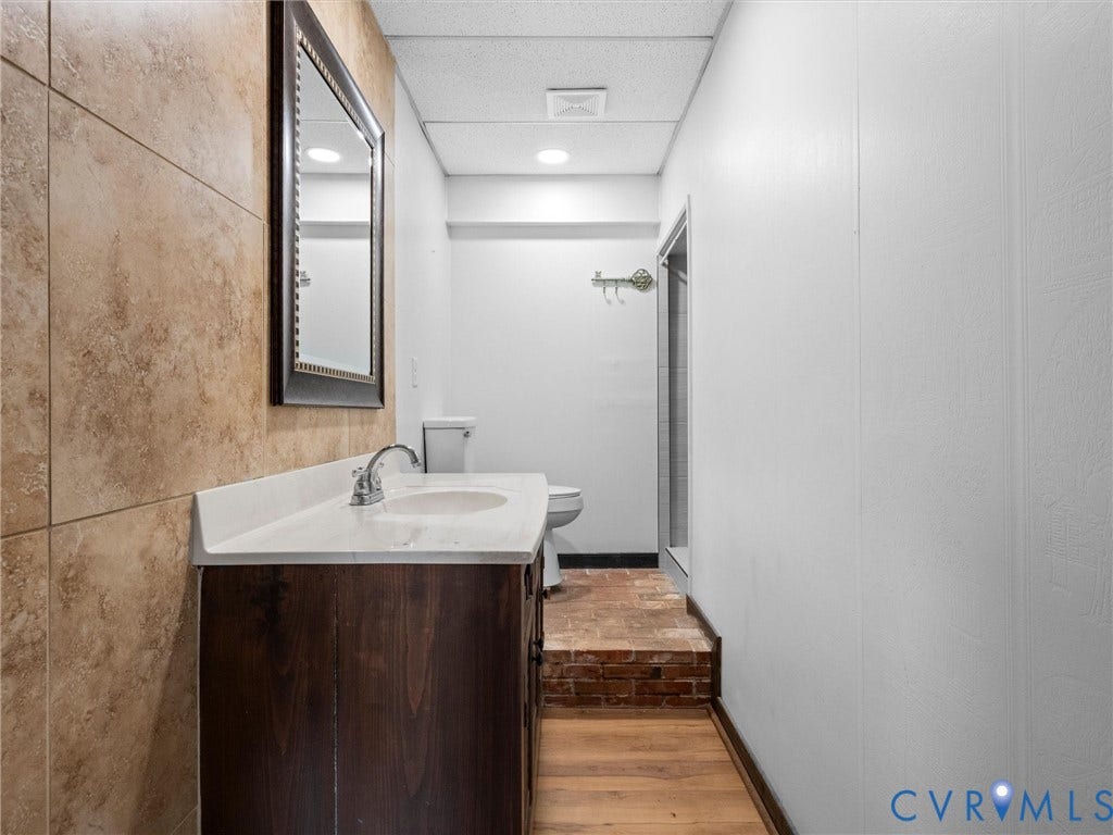 Wood-finish flooring, full vanity with integrated sink, framed mirror, tiled wall, and brick-style flooring