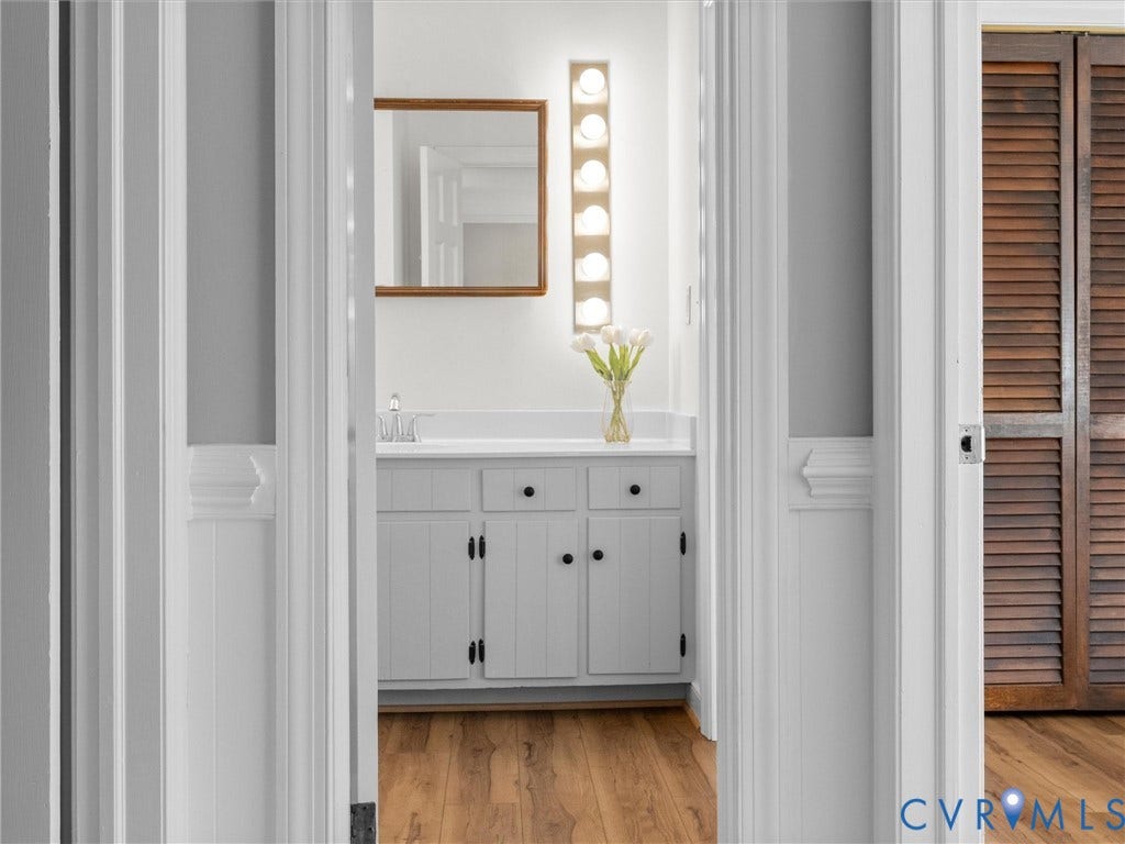 White vanity with integrated sink, wood-finish flooring, wood-framed mirror, and vertical vanity lighting