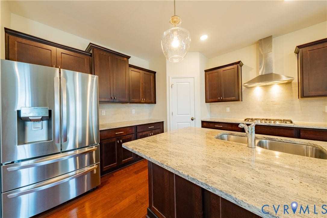 Kitchen featuring wood-finish flooring, dark wood cabinetry, a center island with a double basin sink, stainless steel appliances, and a tiled backsplash