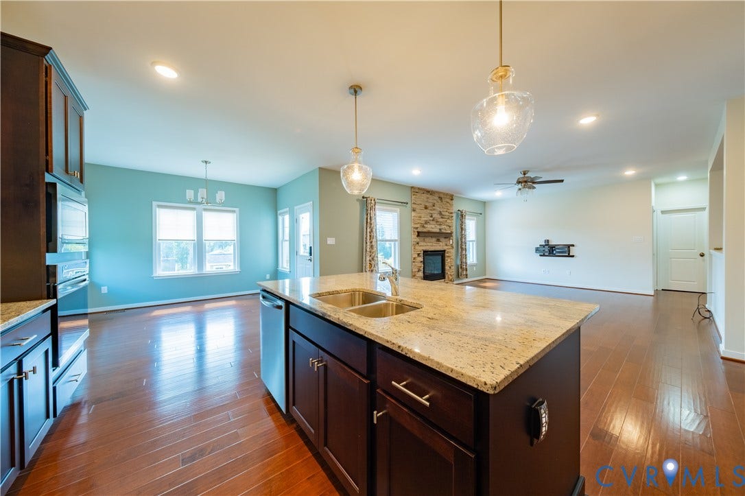 Kitchen island with a double basin sink and granite-finish countertop, wood-finish flooring, recessed lighting, and a stone-clad fireplace