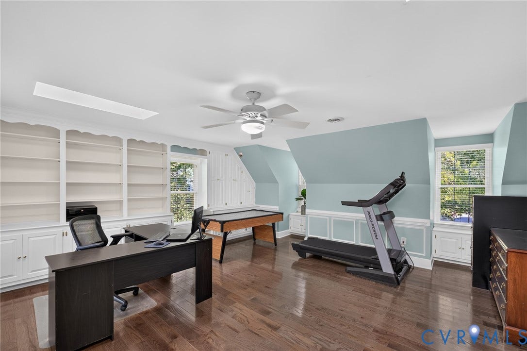 12800 Leffingwell Court, Henrico, VA 23233 Spacious home office featuring hardwood flooring, built-in white cabinetry with open shelving, soft blue accent walls, ceiling fan with integrated lighting, and large windows providing natural illumination throughout the room