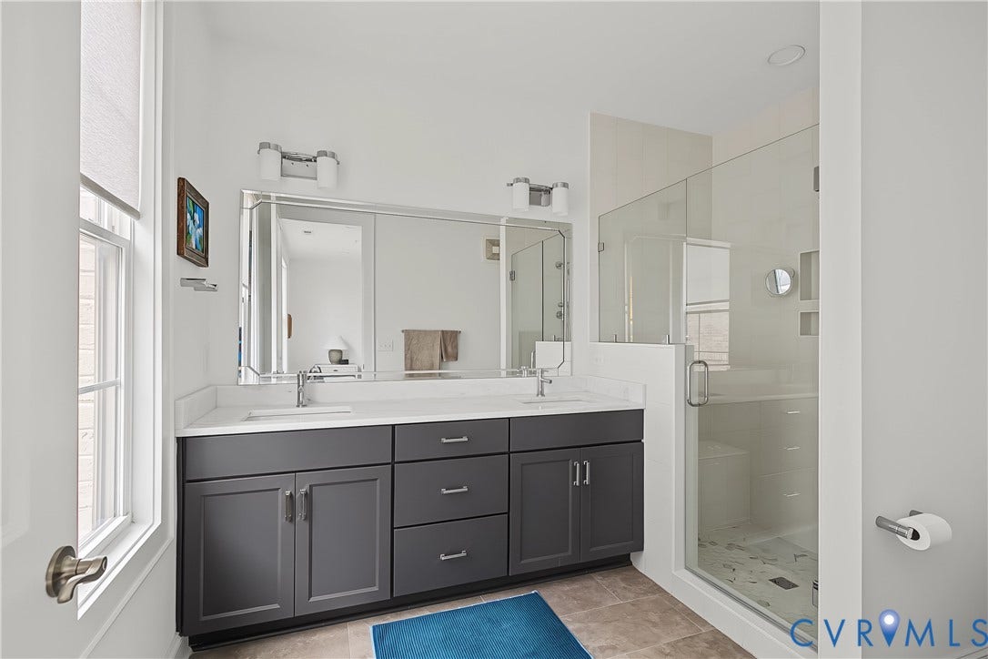 Dual vanity with gray cabinetry and white countertops, large wall-mounted mirror, and a glass-enclosed shower