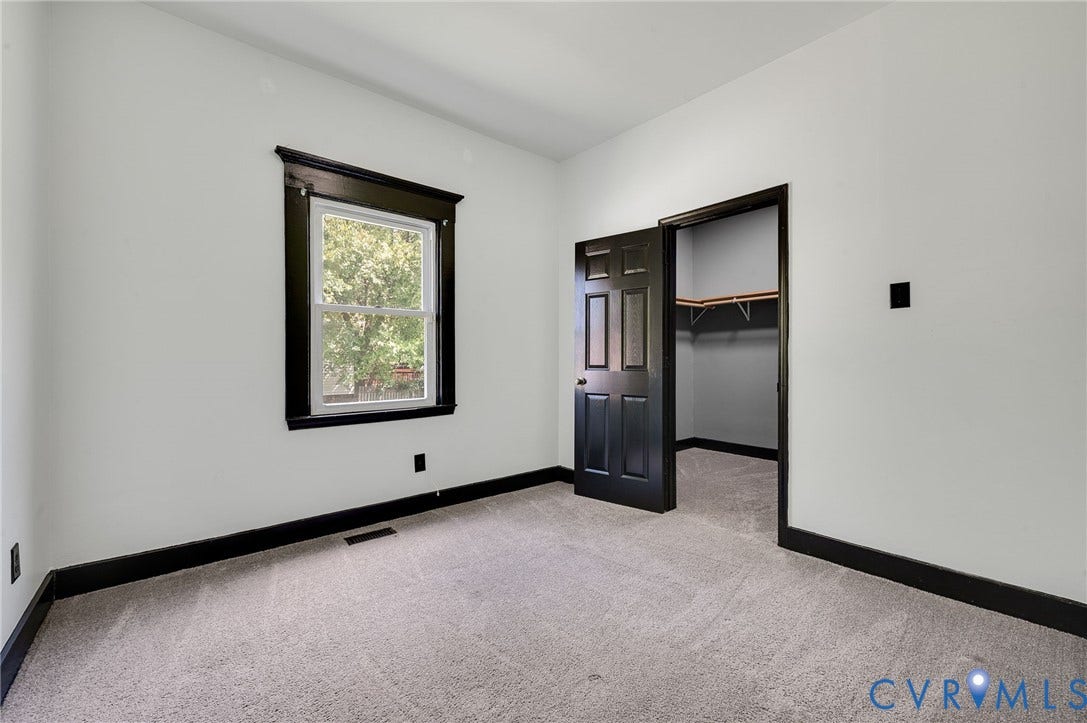 Light gray wall paint, dark trim accents, light gray carpet flooring, window with exterior foliage, and a built-in closet with shelving