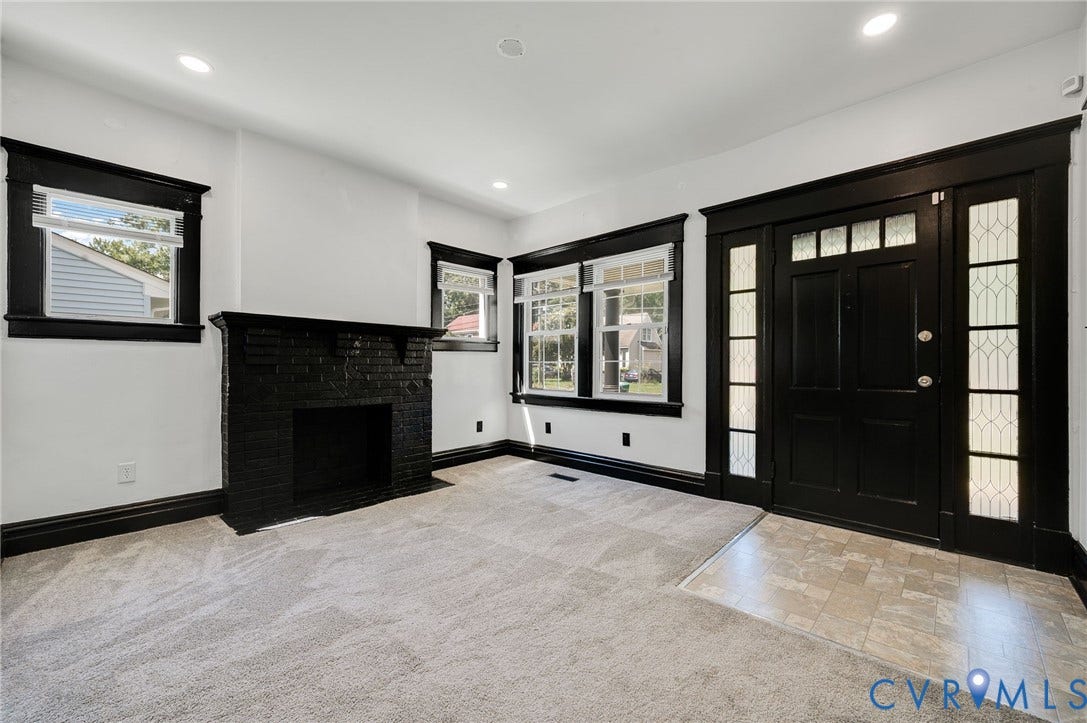 Sophisticated living space featuring a black brick fireplace with full-height surround, recessed ceiling lighting, large multi-pane windows with black trim, black entry door with sidelights, and light wood-finish flooring throughout