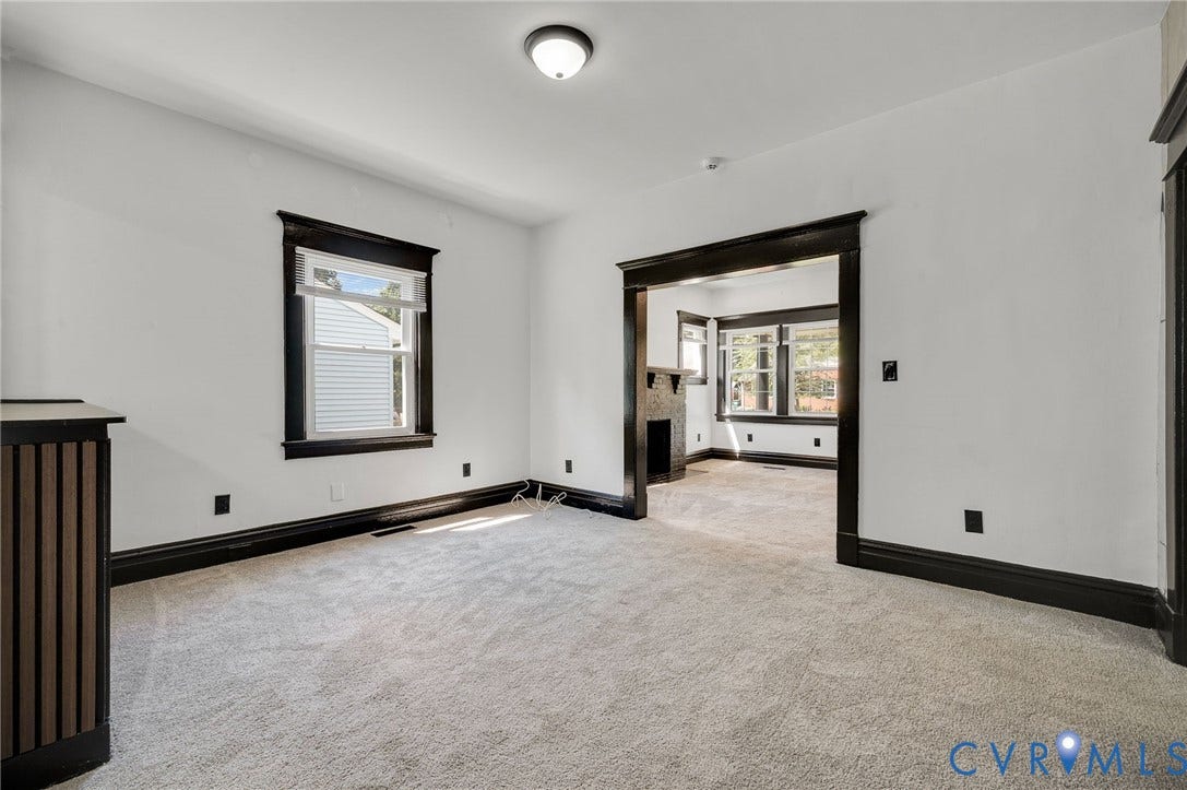 Spacious room featuring neutral carpeting, black-trimmed architectural detailing along the walls, a large window with black frame and interior shutters, and open doorway leading to adjacent spaces