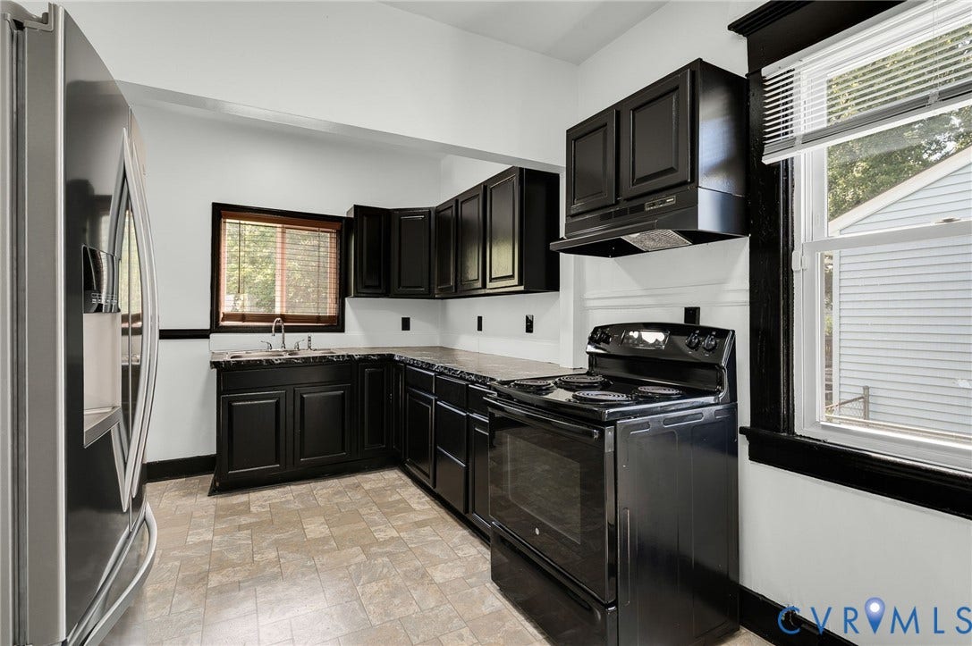 Kitchen featuring black cabinetry, light-toned flooring, dark countertops, a window with natural light, and a stainless steel refrigerator