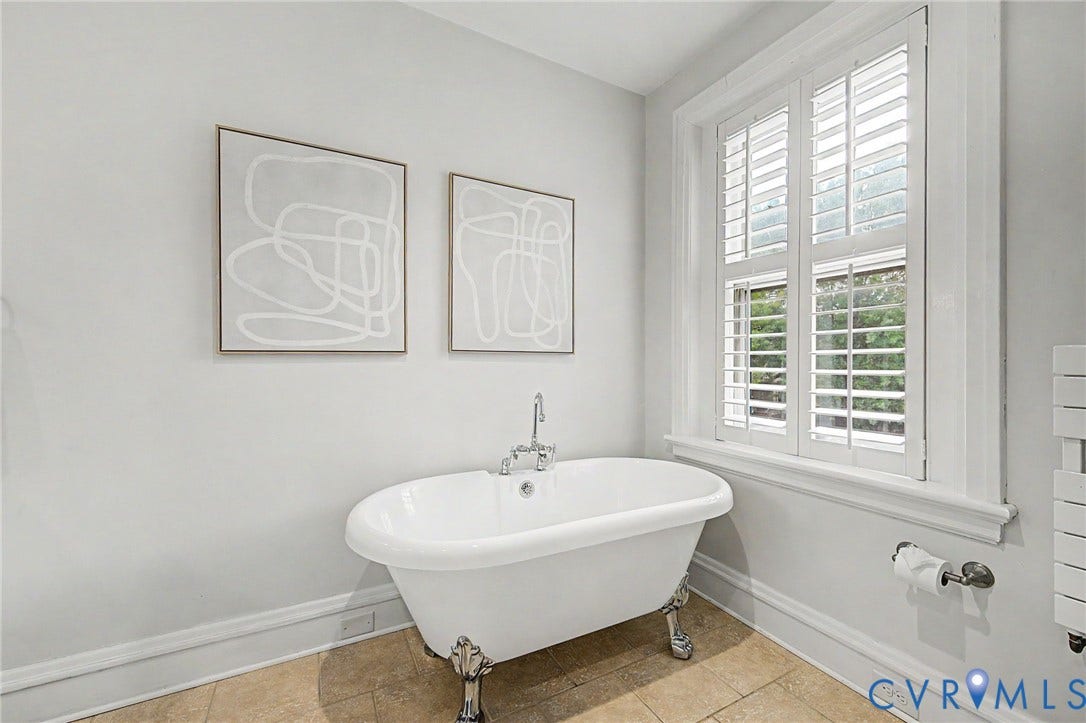Freestanding clawfoot bathtub with chrome fixtures, window with white plantation shutters, and light-toned tile flooring