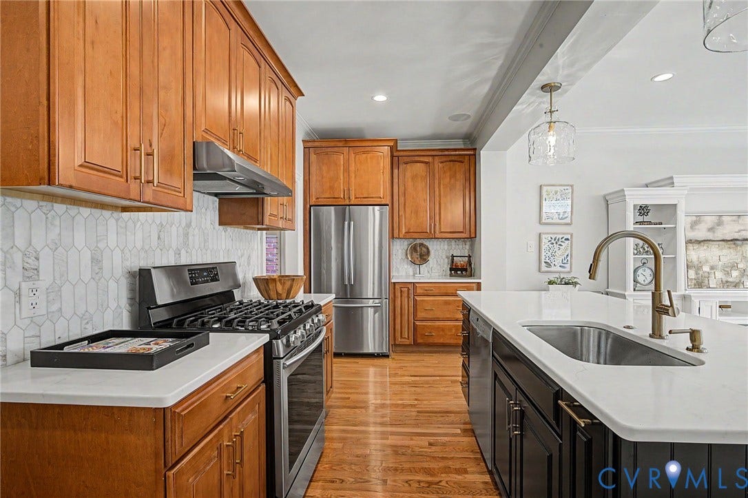 Kitchen featuring wood cabinetry, stainless steel appliances, a white tile backsplash, an island with an undermount sink, and wood-finish flooring
