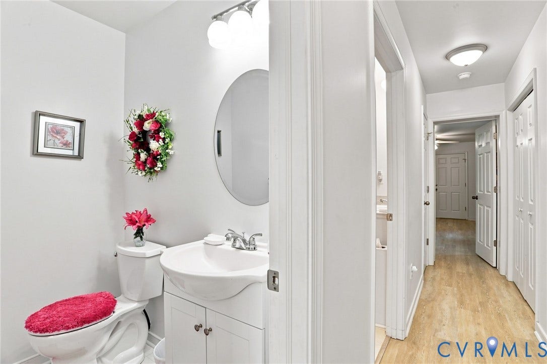 4512 Forrestal Road, Chester, VA 23831 Bright and functional powder room featuring a round mirror, pedestal sink with chrome faucet, white cabinetry with storage, ceramic tile walls, and wood-finish flooring