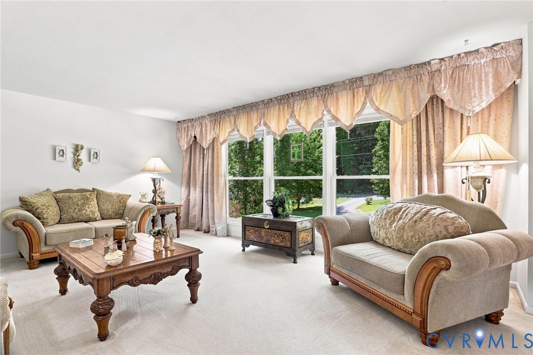 4512 Forrestal Road, Chester, VA 23831 Elegant formal living space featuring expansive multi-pane windows with coordinating drapery, light wood-finish flooring throughout, ornate carved wood coffee table, upholstered seating with rolled arms and wood details, and layered window treatments with swag valances and side panels