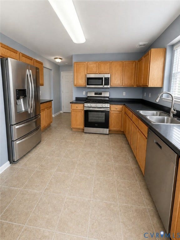Kitchen featuring stainless steel appliances, wood cabinetry, dark countertops, and neutral laminate flooring. Actual.