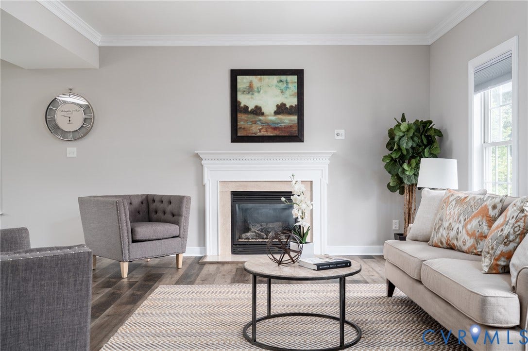 Living area featuring crown molding, a fireplace with a white mantel and stone surround, wood-finish flooring, and a large window providing natural illumination