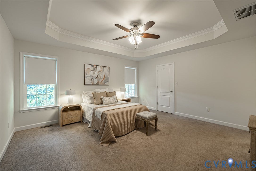 Tray ceiling with crown molding, ceiling fan with light fixture, light-toned carpeting, two windows with roller shades, and a paneled interior door