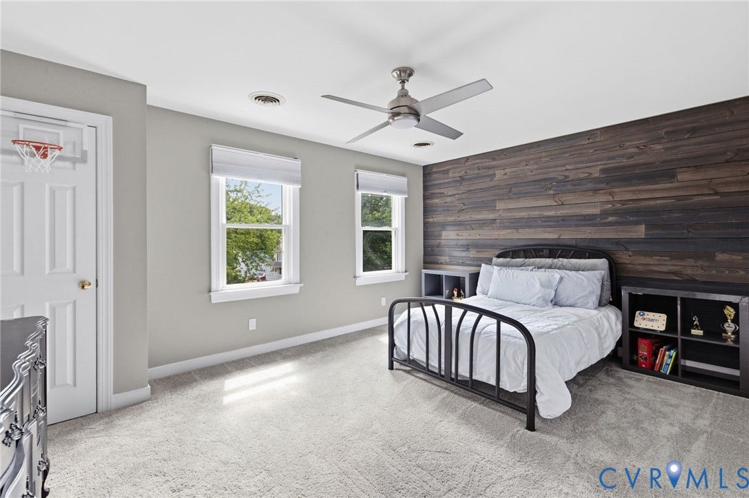 Spacious primary bedroom showcasing a dark wood-finish accent wall, dual casement windows with natural light, ceiling fan, built-in shelving with storage cubbies, and carpeted flooring throughout