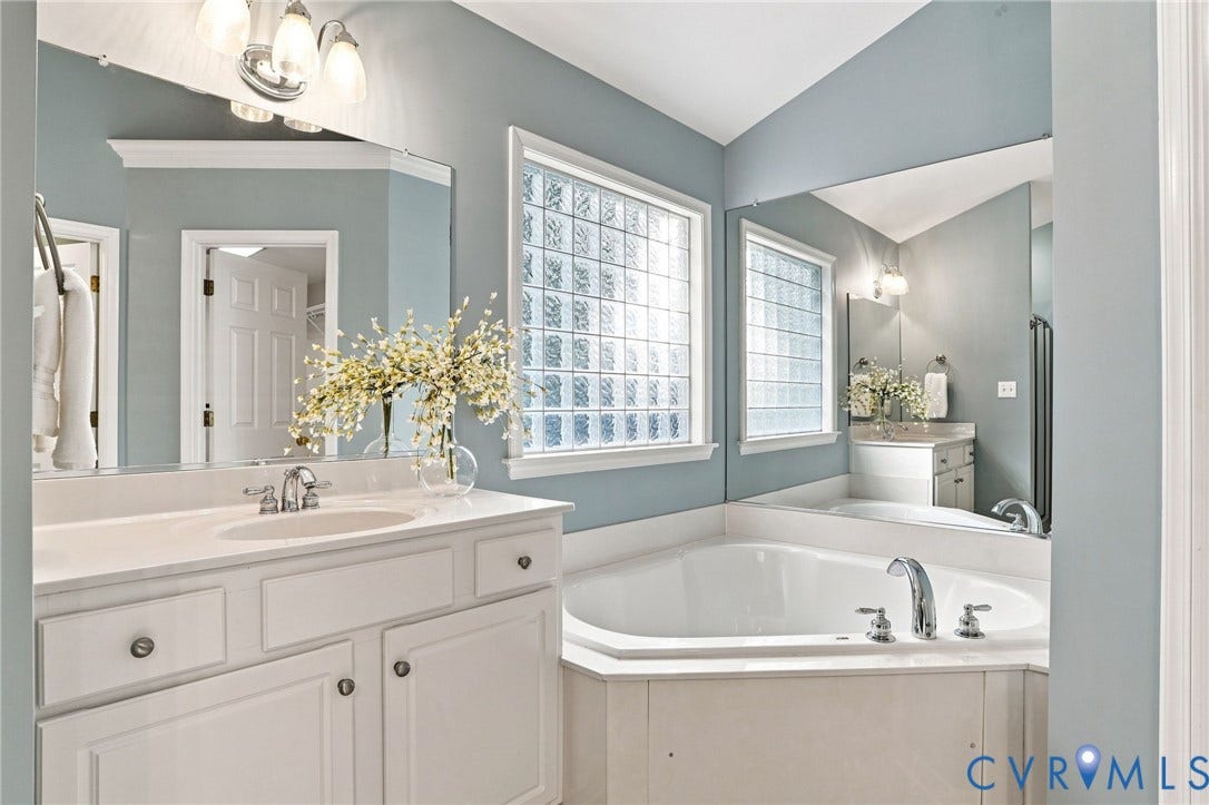 Dual vanity with integrated sinks, corner soaking tub, two privacy glass windows, and chrome fixtures