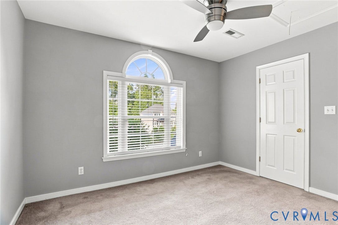 Bright, neutral bedroom featuring neutral carpeting, ceiling fan with integrated lighting, and abundant natural light from a large arched window with plantation shutters