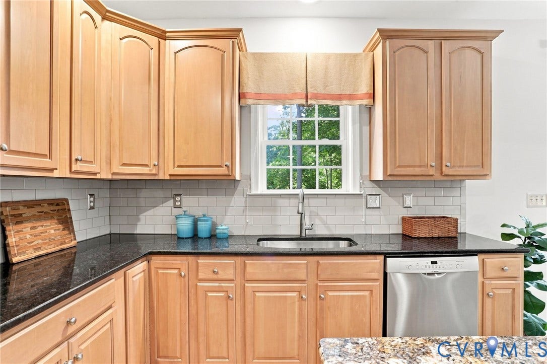 Light wood cabinetry, dark countertops, subway tile backsplash, stainless steel dishwasher, and a stainless steel sink with a gooseneck faucet