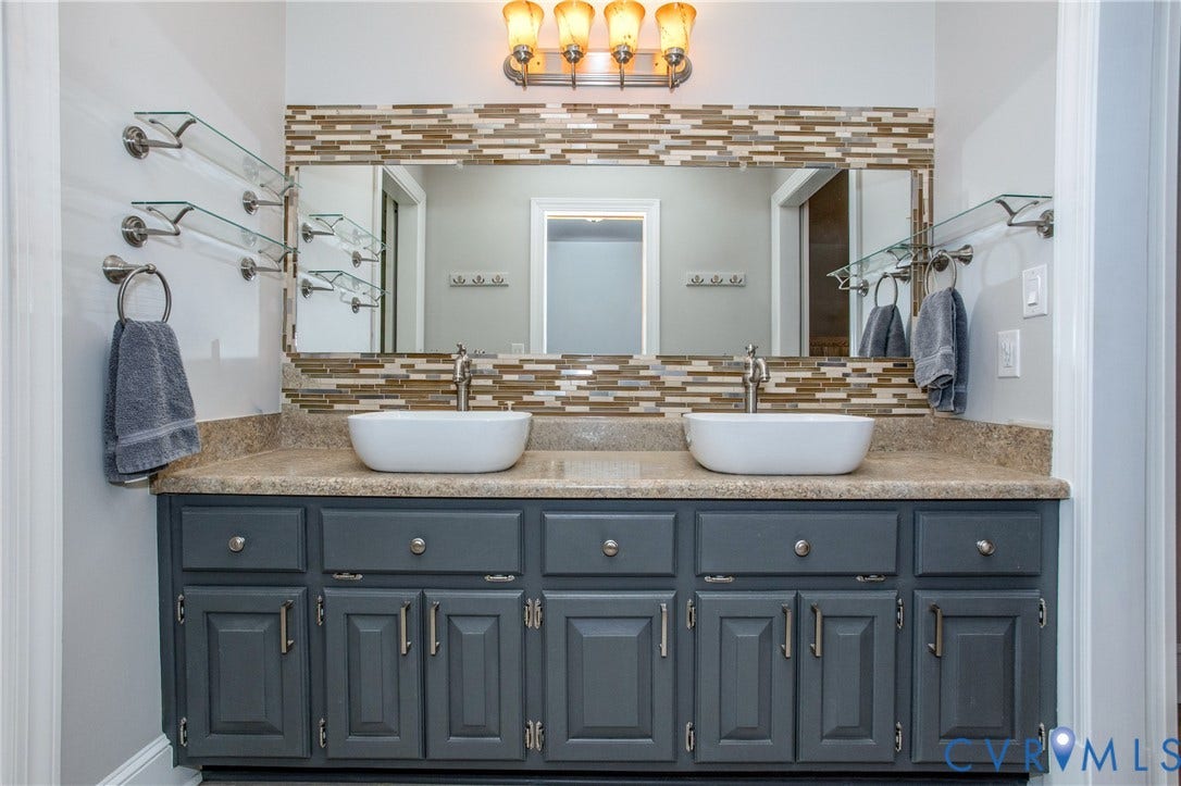 14530 Fox Knoll Drive, South Chesterfield, VA 23834 Dual vessel sinks with individual faucets, ample vanity storage with dark cabinetry, mosaic tile mirror frame, and glass shelving