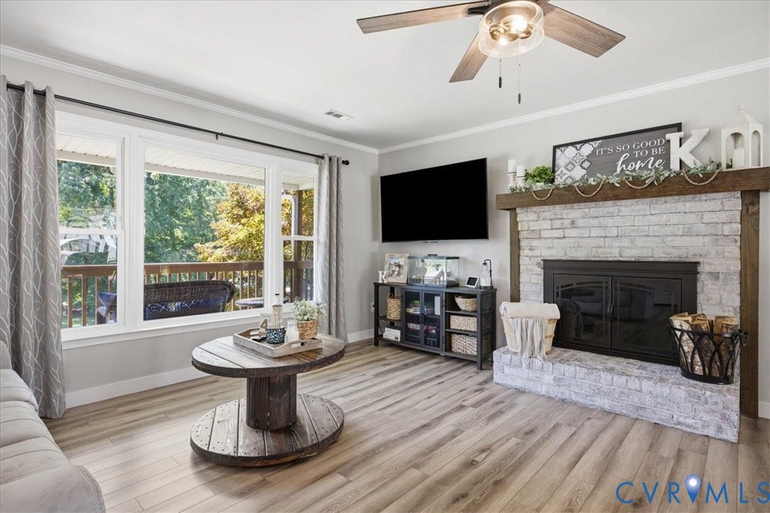 7900 Valencia Road, Chesterfield, VA 23832 Wood-finish flooring throughout, a brick fireplace with a wood mantle, a ceiling fan with integrated lighting, a large window overlooking a deck, and crown molding