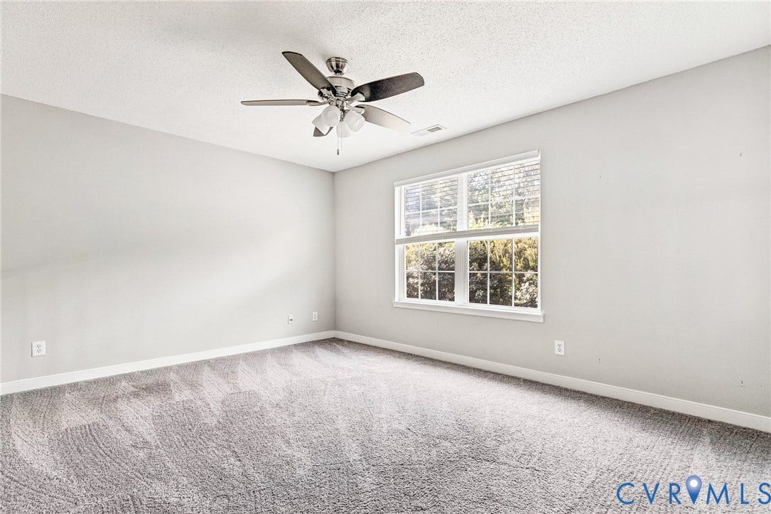 Bright, generously proportioned room with neutral wall finishes, plush carpeting, large multi-pane window with natural light, and brushed nickel ceiling fan with integrated lighting