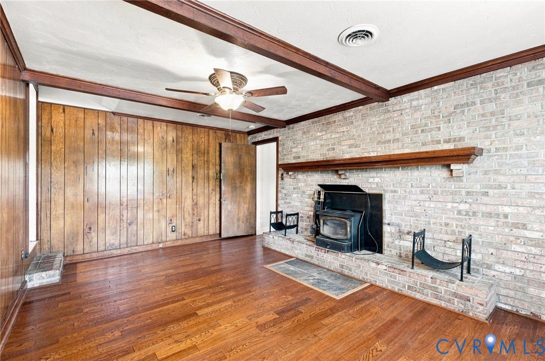 2707 Academy Road, Powhatan, VA 23139 Wood-finish flooring, exposed ceiling beams, wood-paneled wall, brick fireplace with wood mantel, and a ceiling fan with integrated lighting