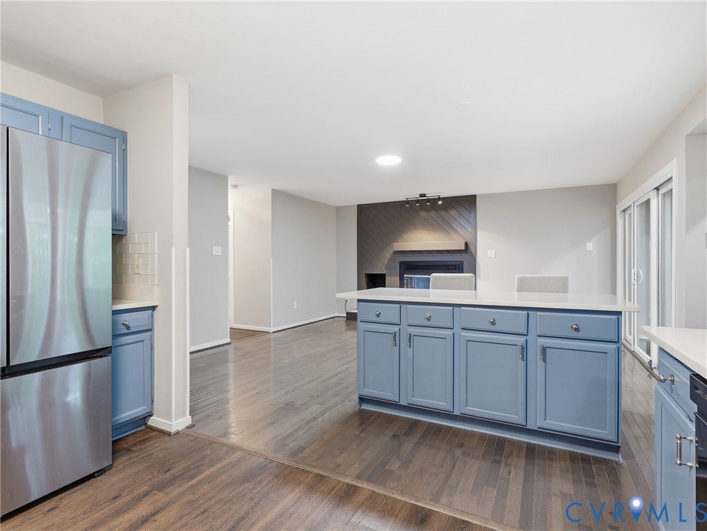 Central island with light-toned countertop, stainless steel refrigerator, wood-finish flooring, and a fireplace with a decorative wood panel surround
