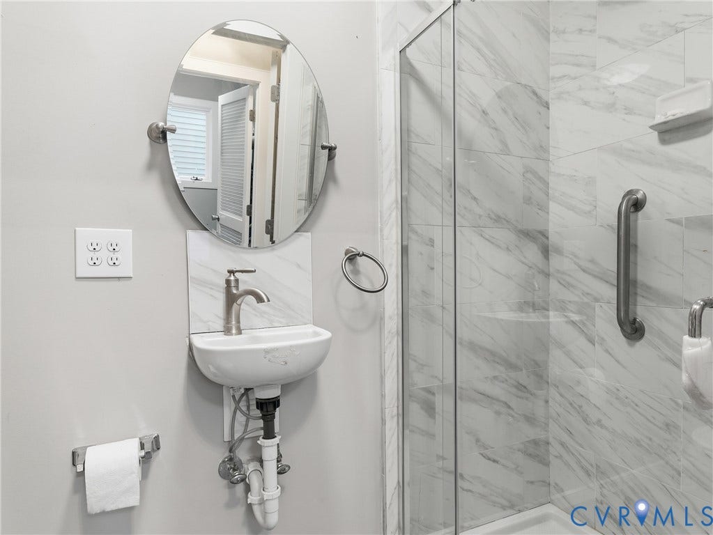 Frameless glass shower enclosure, white porcelain wall-mounted sink, oval vanity mirror, brushed nickel faucet, and marble-patterned shower tile