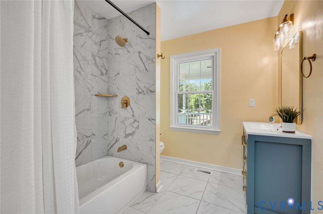 White tile flooring, spacious bathtub with gold-finish fixtures, white vanity with integrated sink, and clear pane window