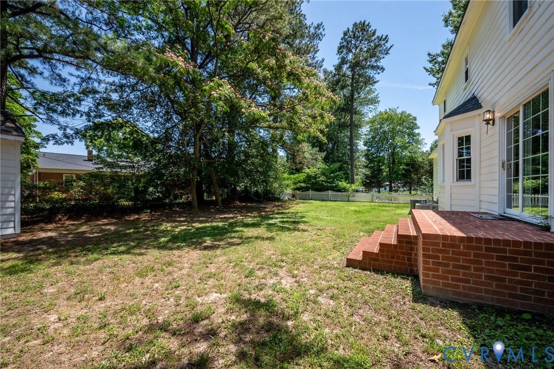 Expansive lawn space with mature trees, a white picket fence, and a brick patio featuring brick steps