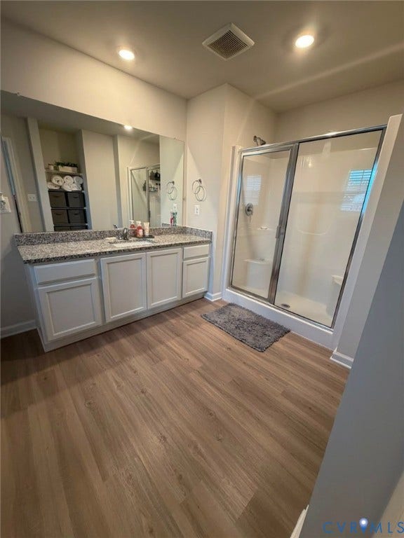 Spacious bathroom featuring wood-finish flooring, a white vanity with panel-front cabinetry, a light-toned countertop, and a glass-enclosed shower