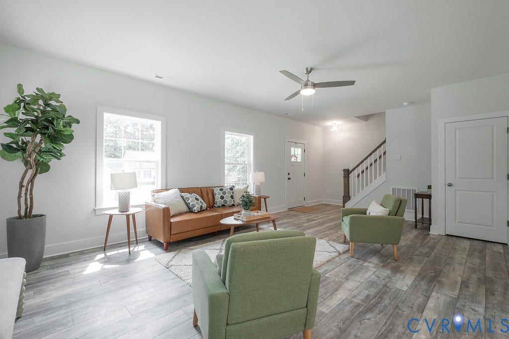 Spacious living area featuring wood-finish flooring, abundant natural light from multiple windows, a ceiling fan with integrated lighting, and a prominent staircase with white risers and dark wood handrails