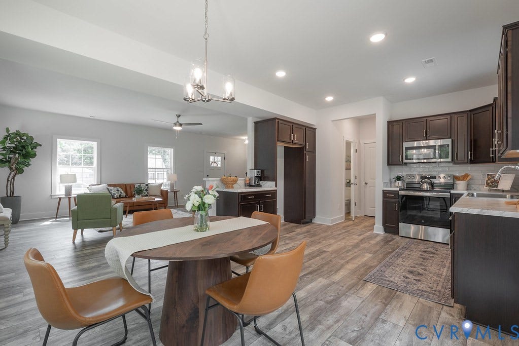 Open-concept living area with wood-finish flooring, a kitchen featuring dark cabinetry, stainless steel appliances, and a tile backsplash, and a dining area with a contemporary chandelier