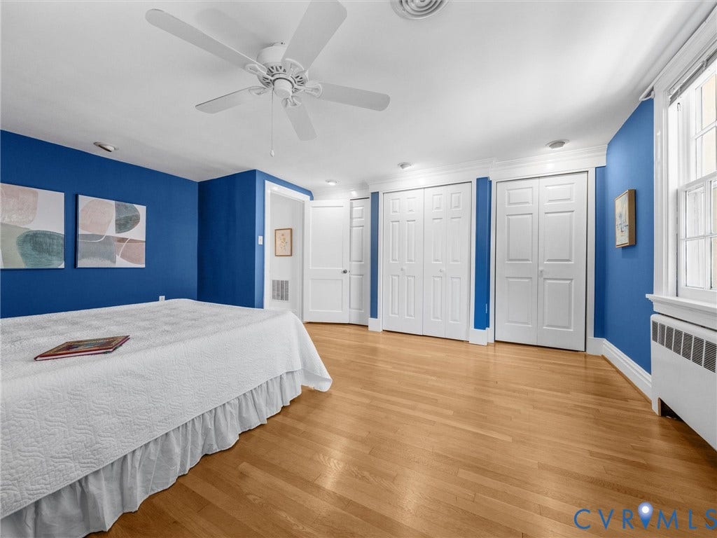 Wood-finish flooring, white trim, multiple closet doors, window with a radiator below, and a ceiling fan