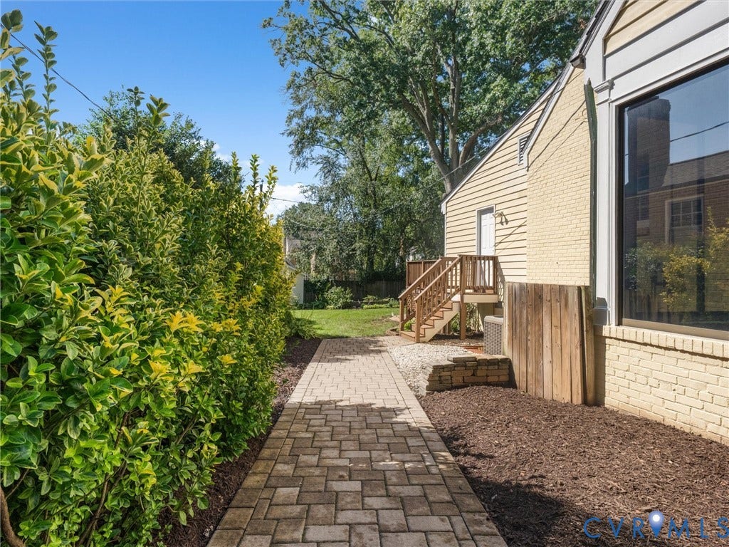 Brick paver pathway with manicured perimeter hedging, leading to a wood deck with stairs and a side entry