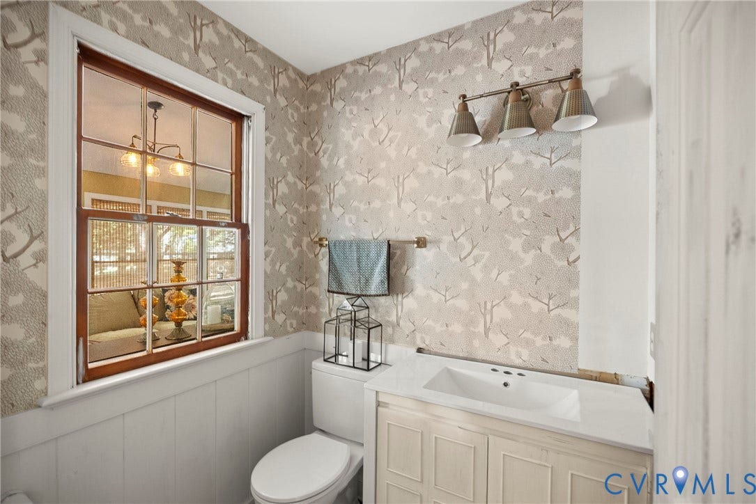 Window with wood-frame sashes, wainscoting, integrated sink with vanity, wall-mounted lighting fixture, and patterned wallpaper