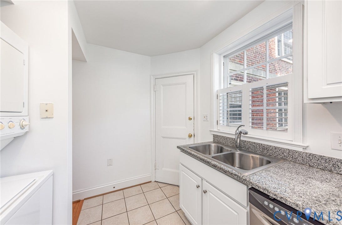 24 W Locke Lane, #1, Richmond, VA 23226 Kitchen area featuring a stainless steel double sink, gray speckled countertop, white cabinetry with silver hardware, and a white window with integrated shutters