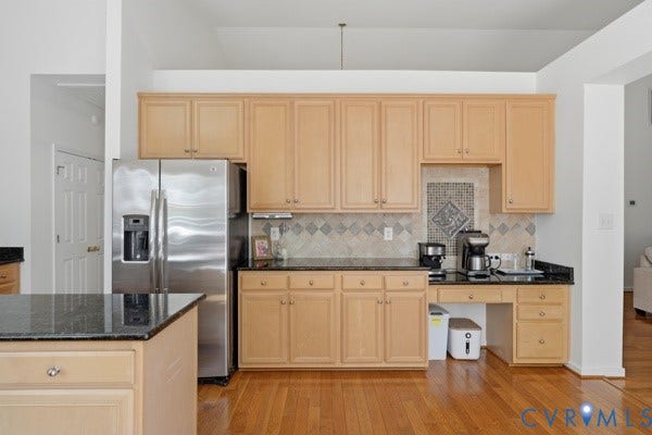 Kitchen featuring wood-finish flooring, light wood cabinetry, stainless steel refrigerator, dark stone countertops, and a decorative tile backsplash